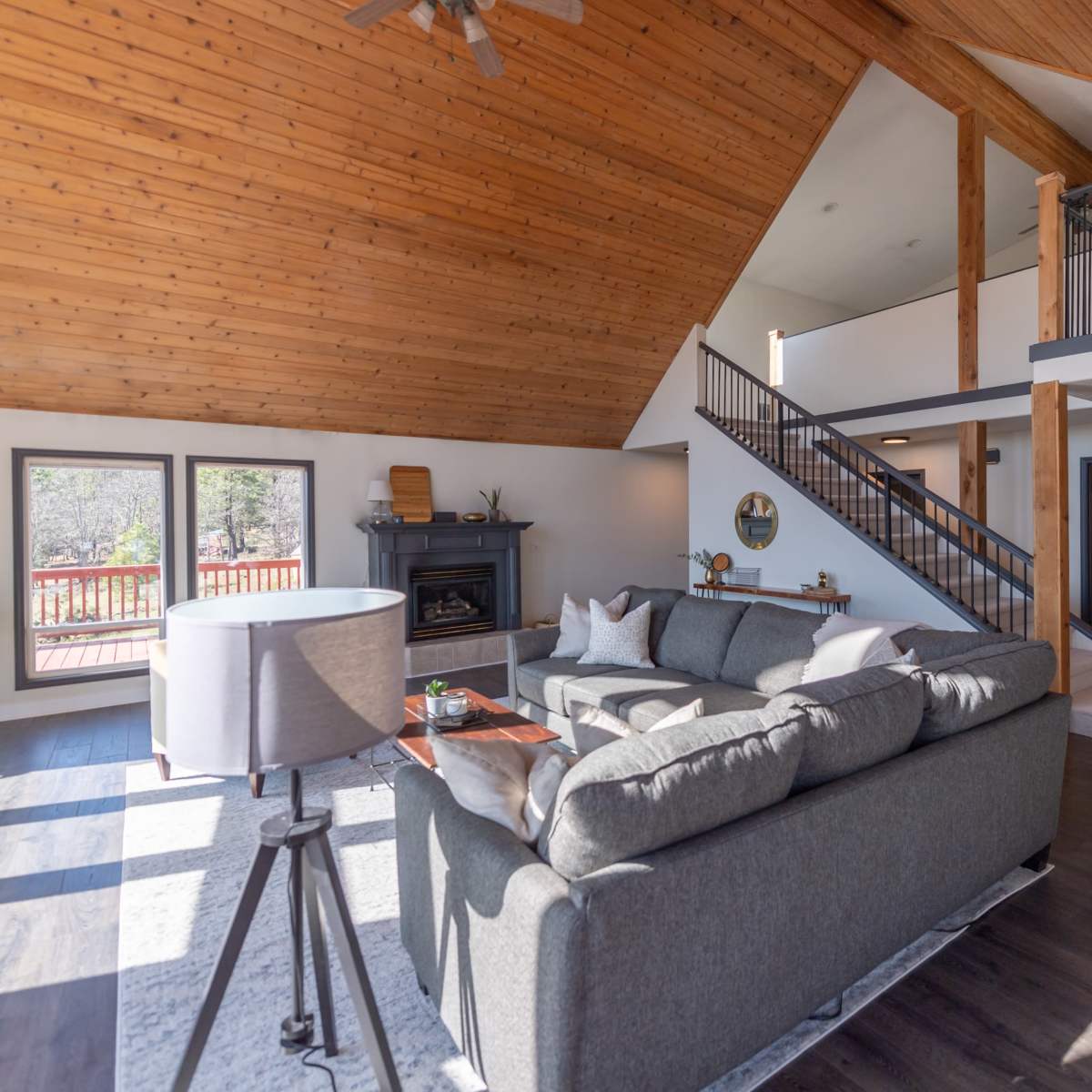 The living room features a spacious layout with a high wooden ceiling, large windows, a gray sectional sofa, and a staircase leading to an upper level.