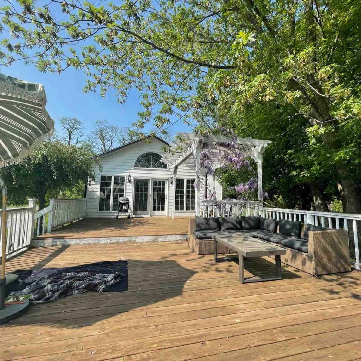 A wooden deck features outdoor seating and a large umbrella, surrounded by lush greenery and a white house with multiple windows.