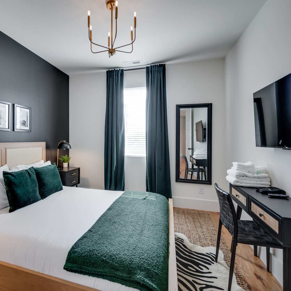 A modern bedroom featuring a bed with a green throw blanket, two black nightstands with lamps, a large mirror, and a television mounted on the wall.