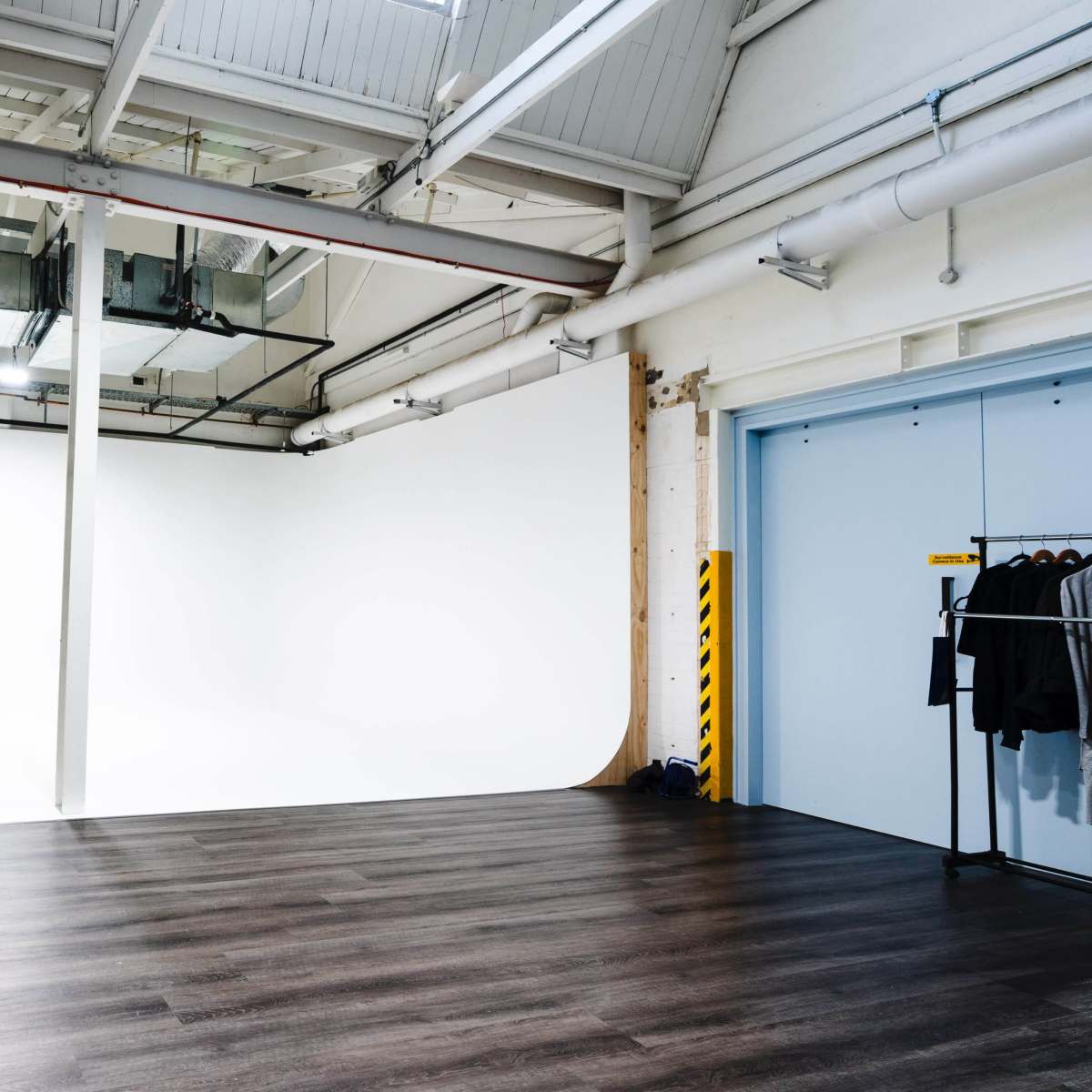 A large, empty studio space with a white backdrop, two black stools, and a clothing rack against a wall.