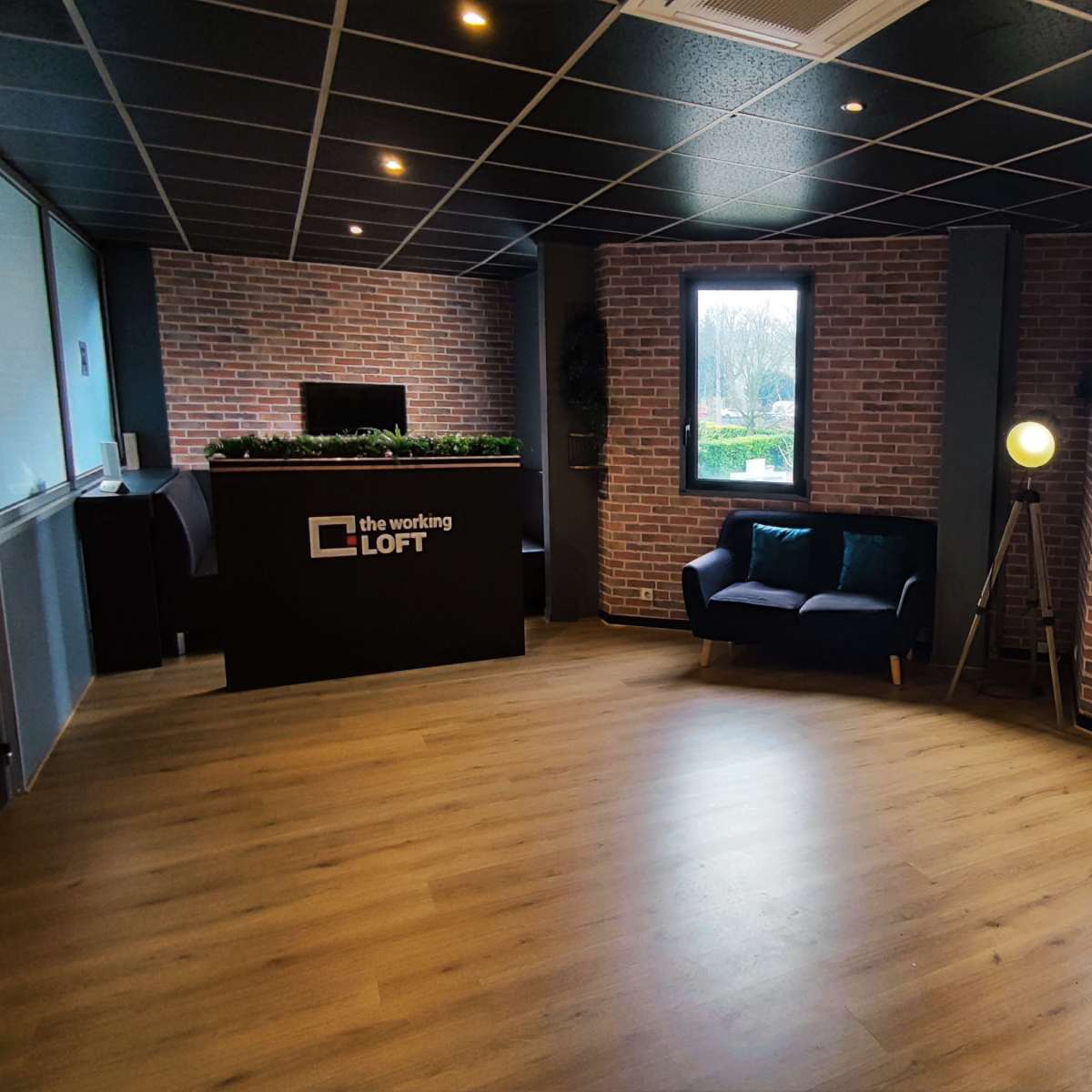 A modern office reception area featuring a brick wall, wooden flooring, a seating area, and a small desk with decorative plants.