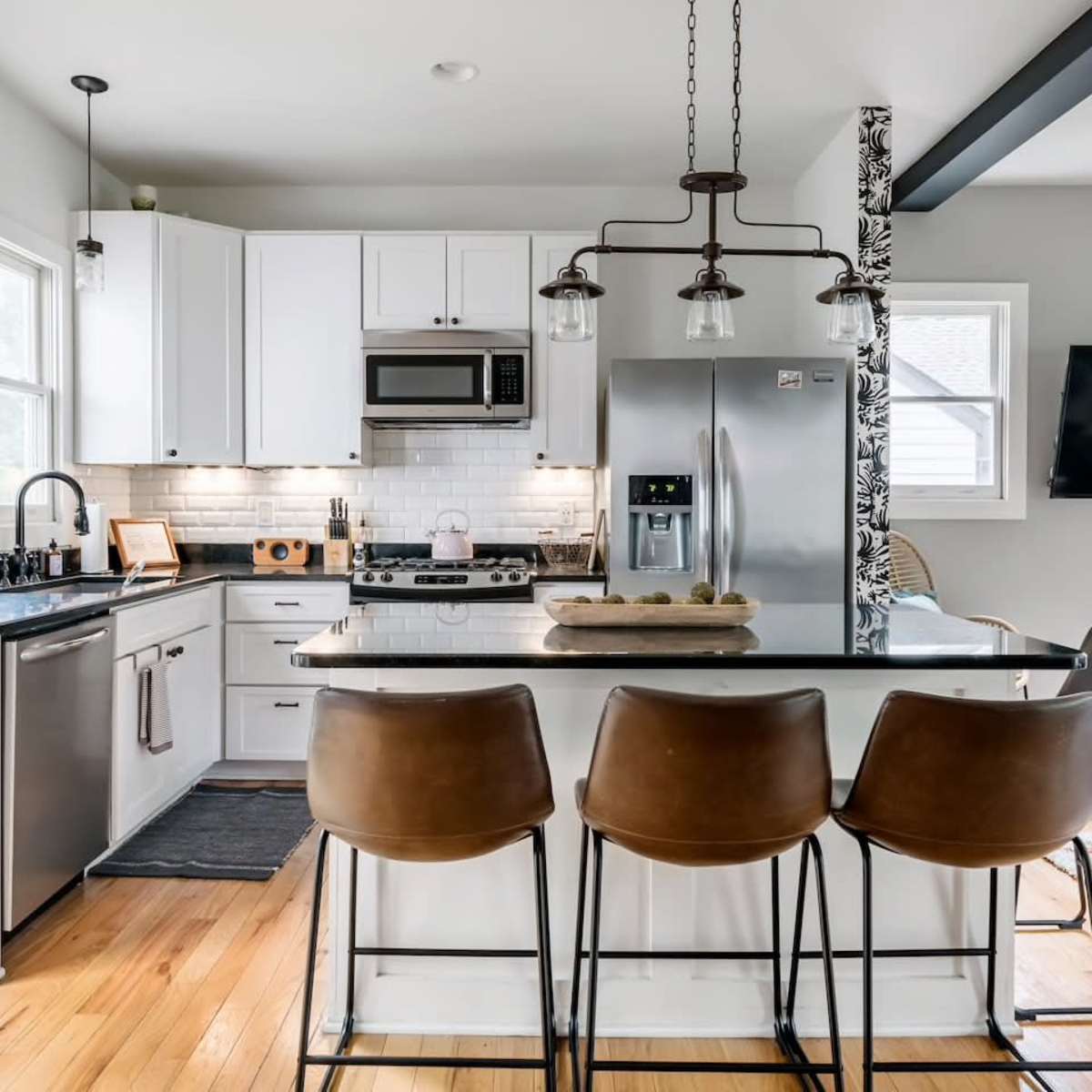 A modern kitchen with white cabinetry, stainless steel appliances, a large island with four brown bar stools, and a television mounted on the wall.