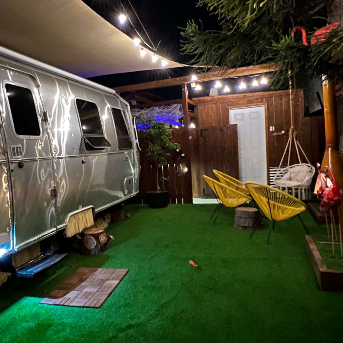 A silver Airstream trailer parked on grass surrounded by string lights, a wooden fence, and two yellow chairs.