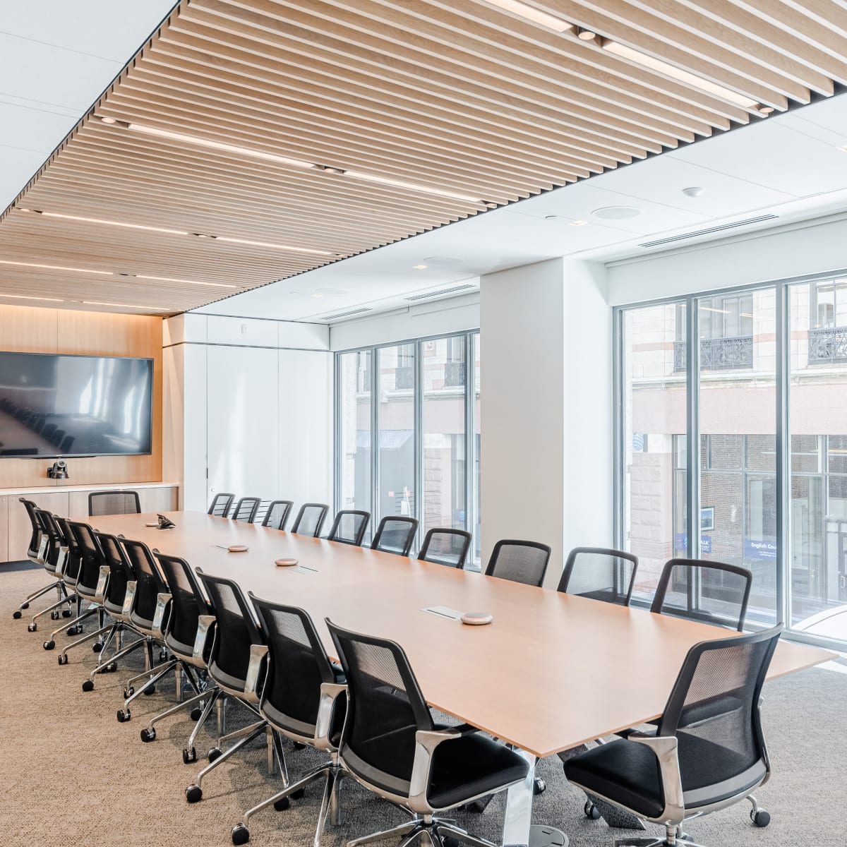 A long conference table with black chairs is set up in a modern meeting room featuring large windows and a wall-mounted screen.