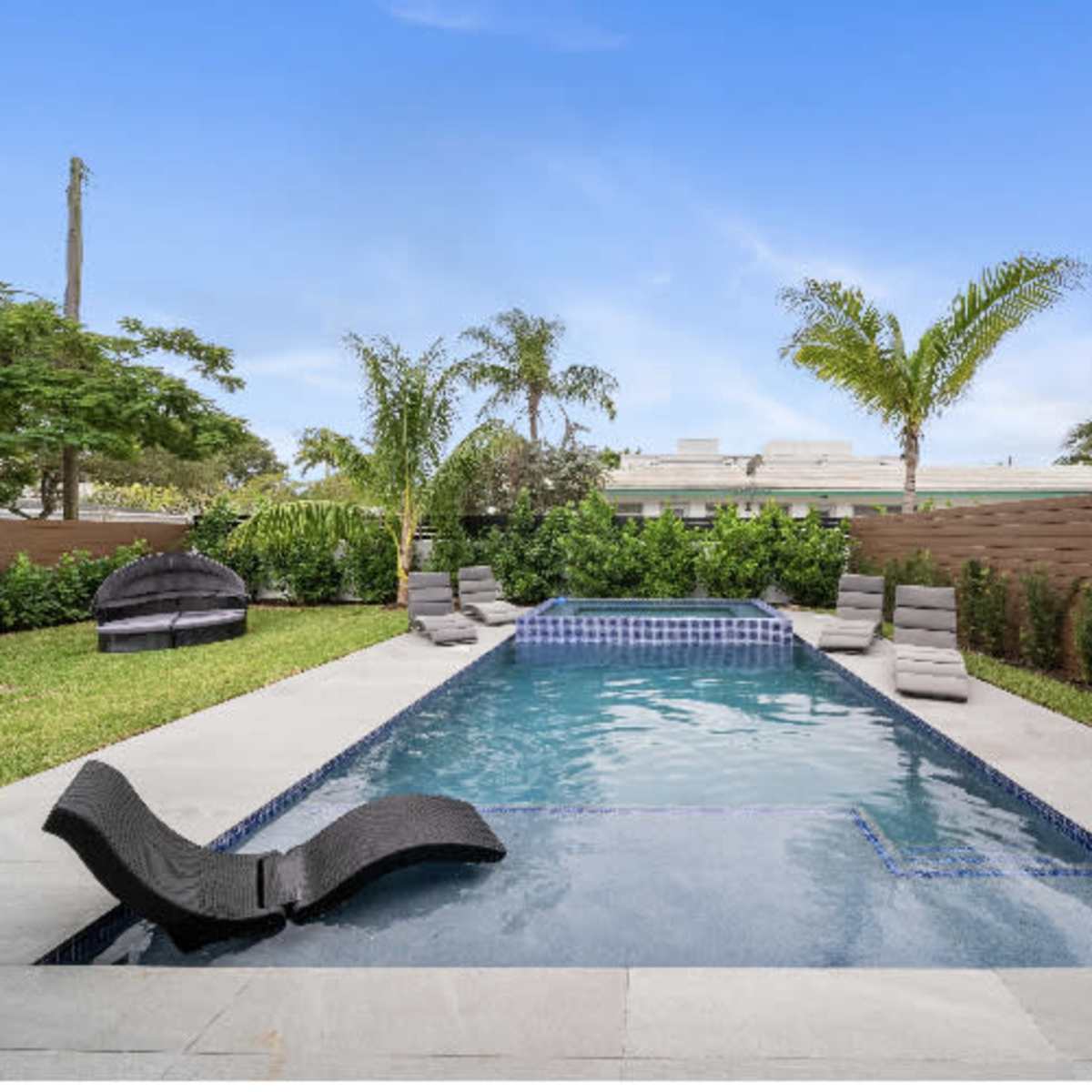 A rectangular swimming pool surrounded by lounge chairs and palm trees, enclosed by a low wall and greenery.