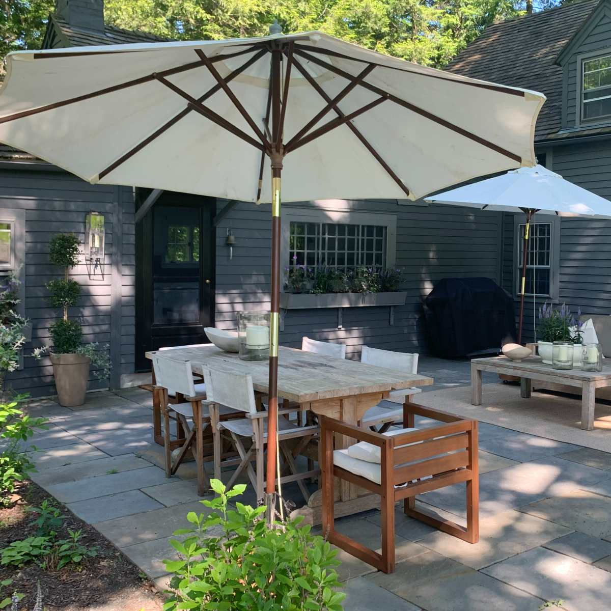 A stone patio area with a wooden dining table and chairs, two large umbrellas, and a lounge area featuring a sofa and coffee table beside a house with gray siding.