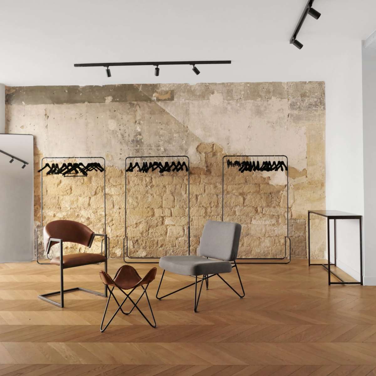 A minimalistic clothing store with two chairs, hanging clothing racks, and a light wooden floor against a textured wall.