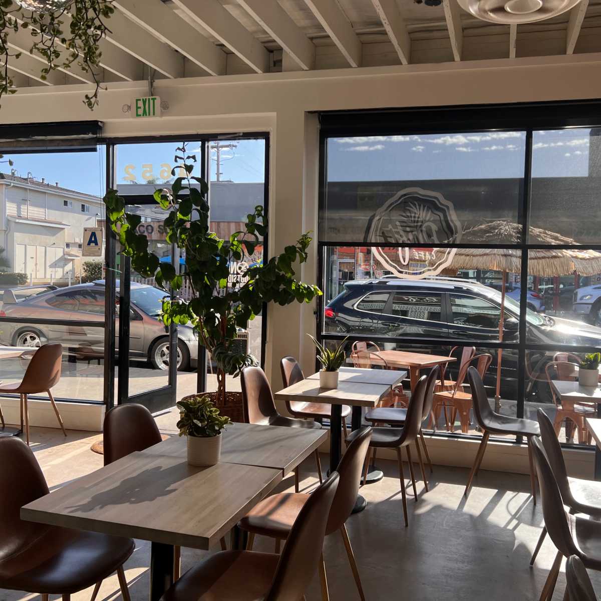 The interior of a cafe features wooden tables and chairs, with large windows allowing natural light and a view of parked cars outside.