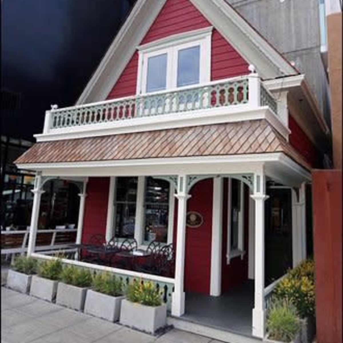 A red, two-story house with a steep roof, decorative balcony, and a paved walkway surrounded by planters.