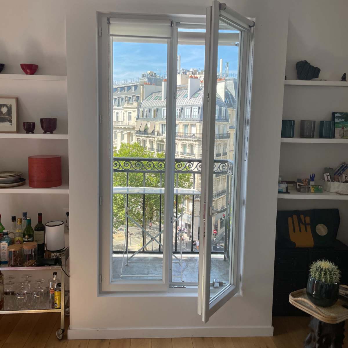 A white window with open panels frames a view of buildings and trees outside, set within a room that has shelves displaying various objects.