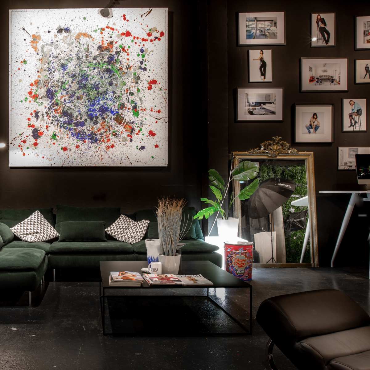 A modern interior with a green couch, a coffee table with magazines, and a large abstract painting on the wall, surrounded by various framed photographs and plants.