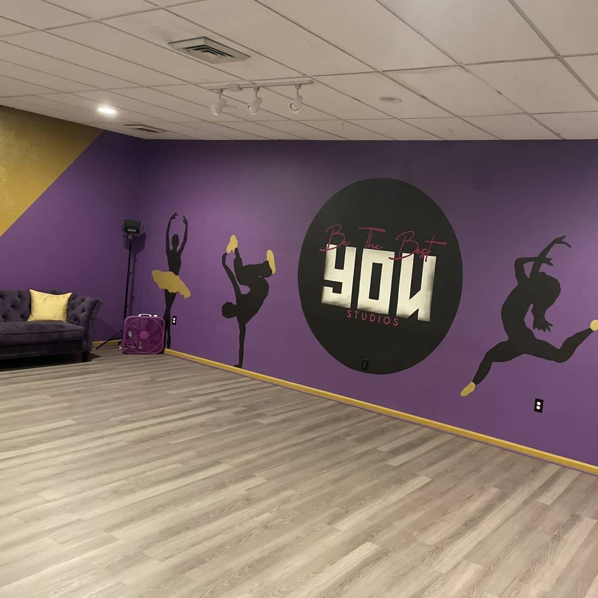 A dance studio with a purple and yellow wall featuring silhouettes of dancers and the words "Be It But YOU Studios."