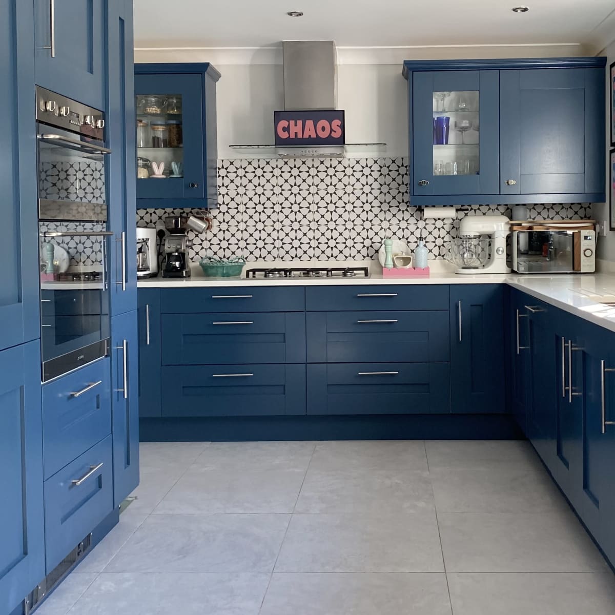 A modern kitchen features blue cabinets, white countertops, and a patterned backsplash, with a sign reading "CHAOS" prominently displayed.
