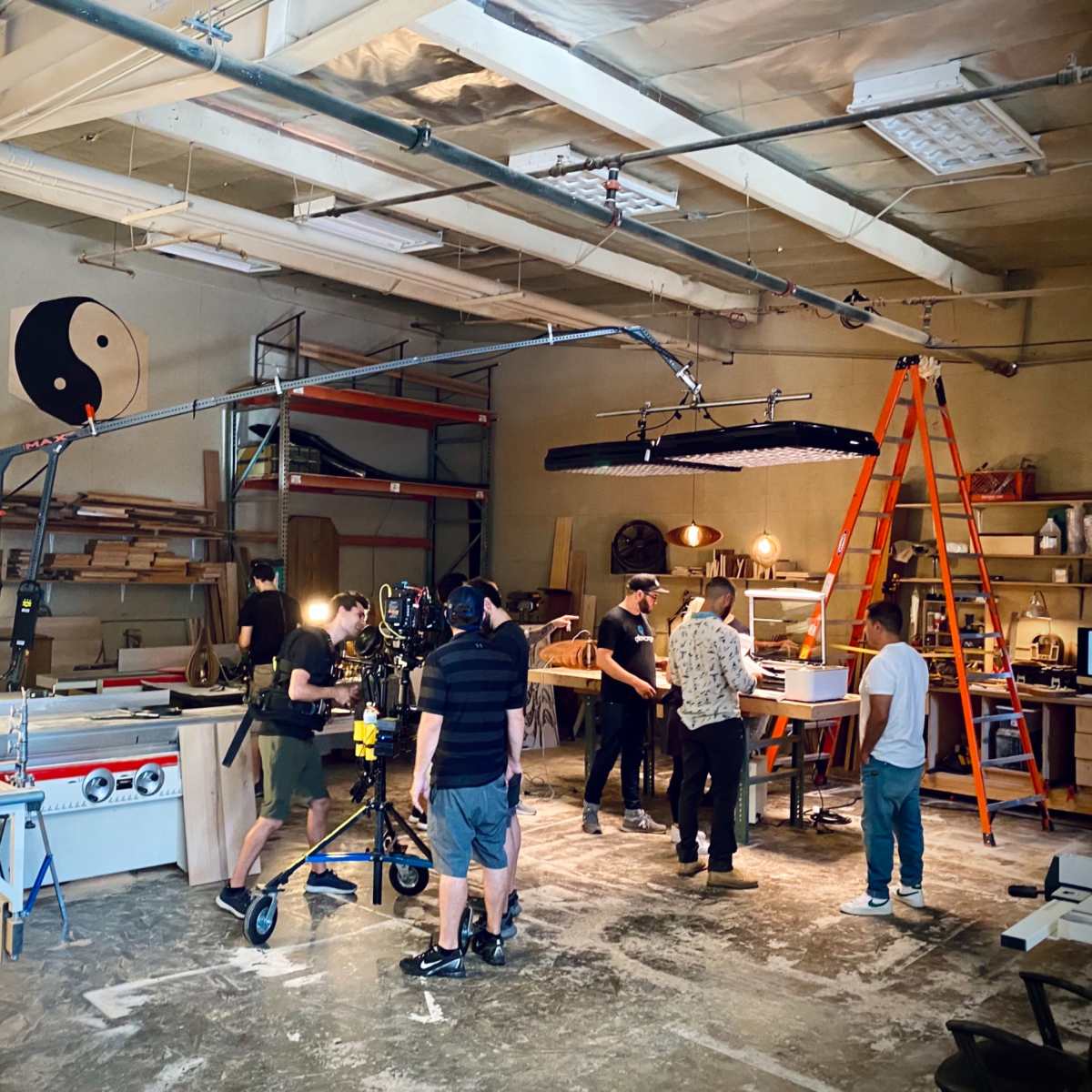A film crew is set up in a workshop, preparing equipment for a shoot, with various tools and furniture visible in the background.