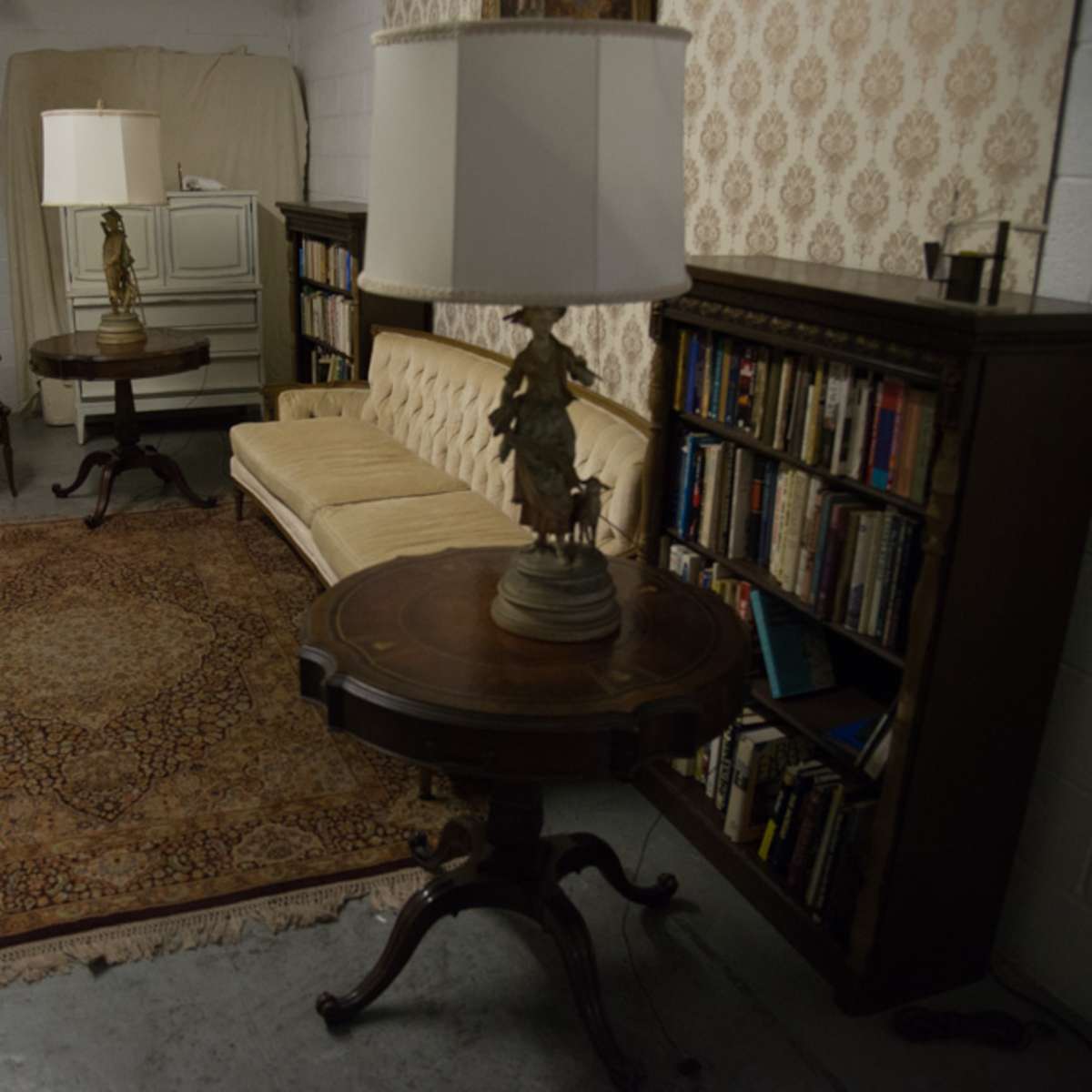 The scene features a cozy sitting area with a vintage sofa, two armchairs, a rug, a lamp on a round table, and a bookshelf filled with books.