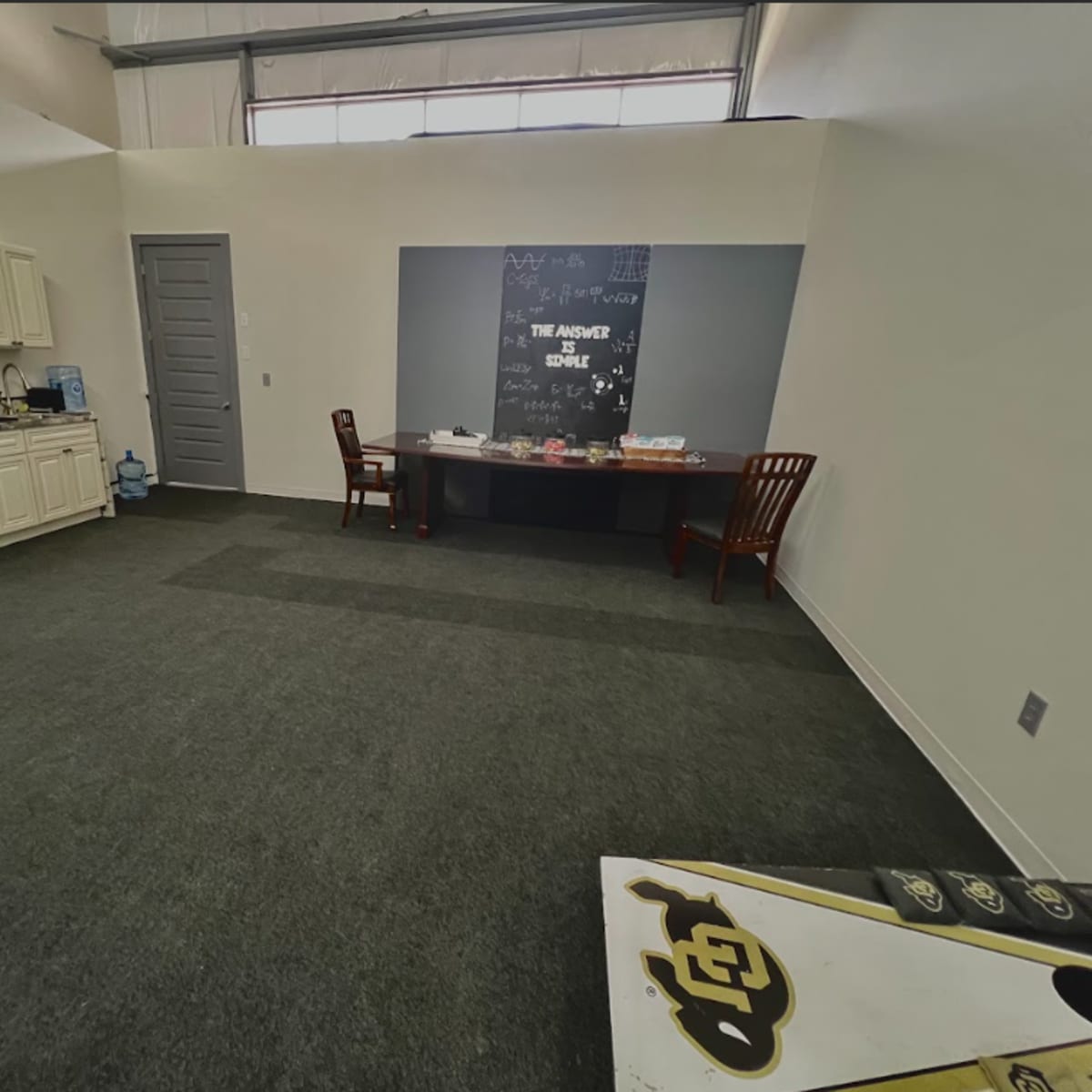 A sparsely furnished room with a kitchenette, two wooden chairs, and a table against a wall featuring a chalkboard.