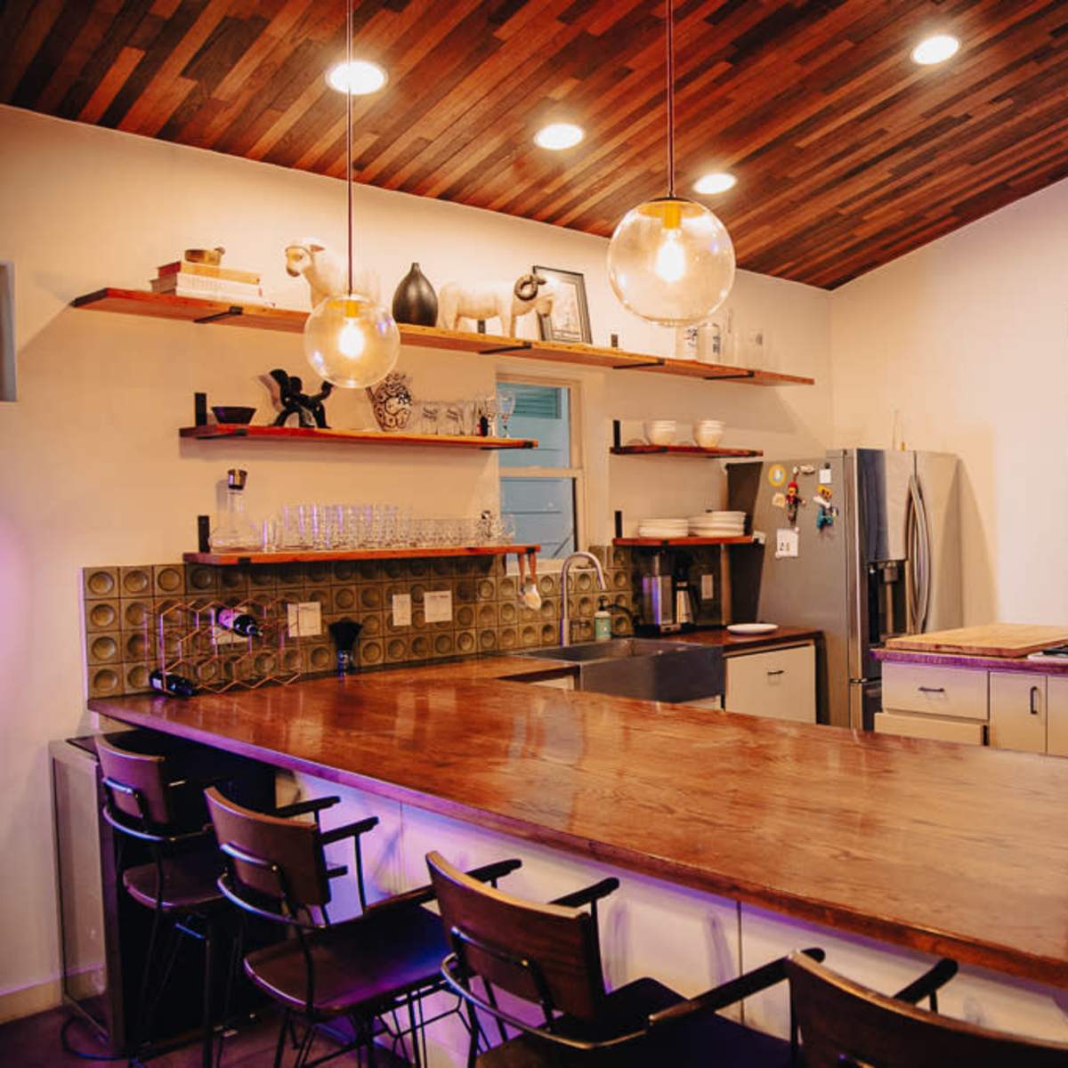 A modern kitchen features a large wooden counter, shelves with kitchenware, and a stainless steel refrigerator, with a view of the outdoors through a wide open window.
