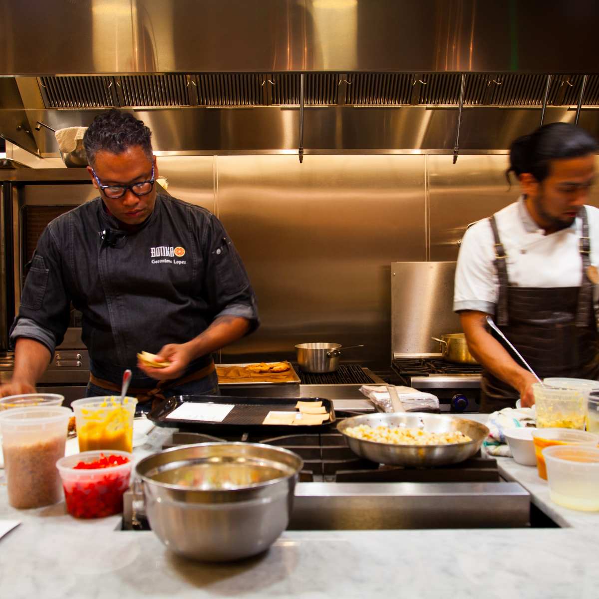 Two chefs are preparing food in a modern kitchen, surrounded by various containers and utensils on a marble countertop.