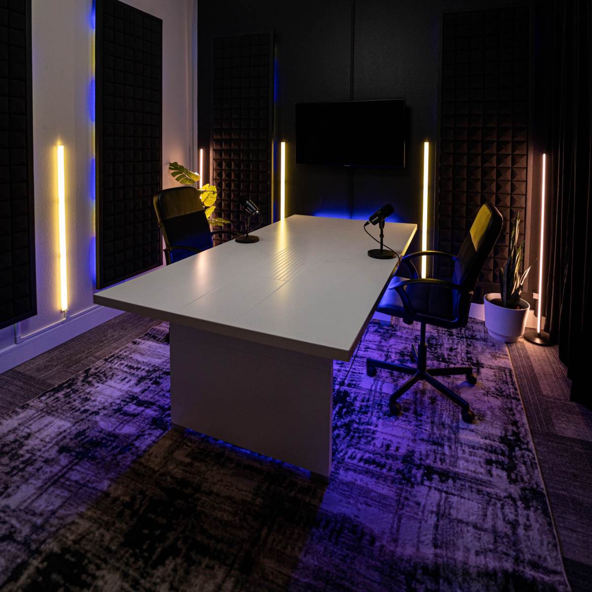 A modern conference room features a central table with microphones, surrounded by two chairs, and illuminated by soft lighting with acoustic panels on the walls.