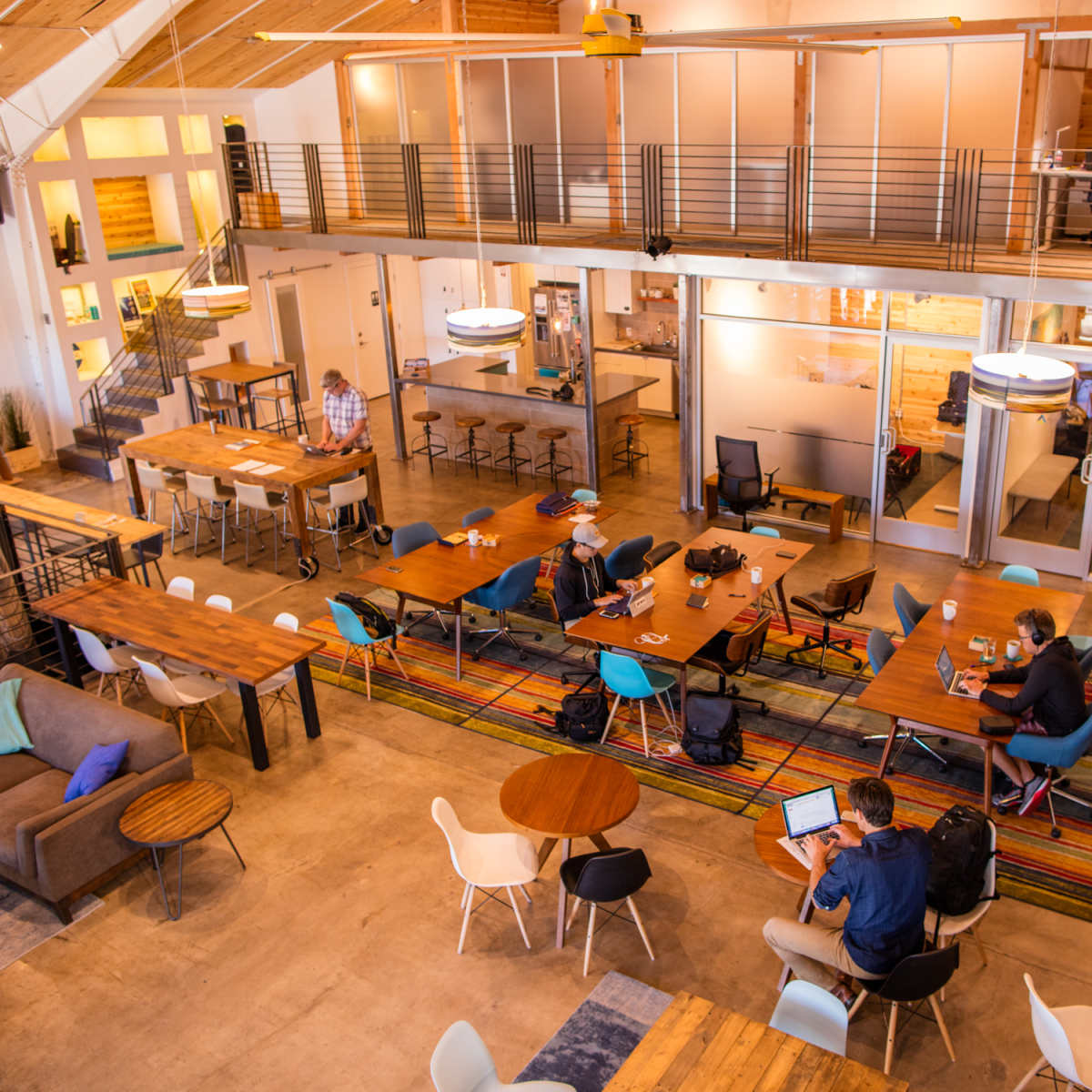 A spacious co-working space features multiple tables, a lounge area, and individuals working on laptops.