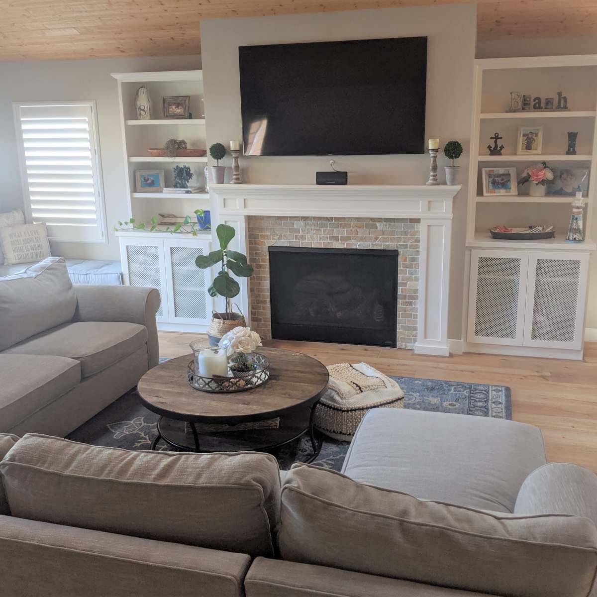 A living room with two gray sofas facing a circular coffee table, a stone fireplace beneath a mounted television, and built-in shelves displaying various items.