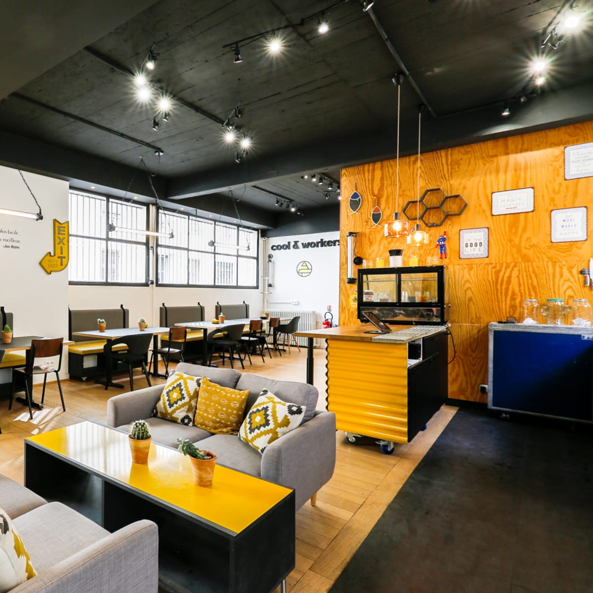 A modern café with wooden seating, tables, potted plants, and yellow accents, illuminated by overhead lights.