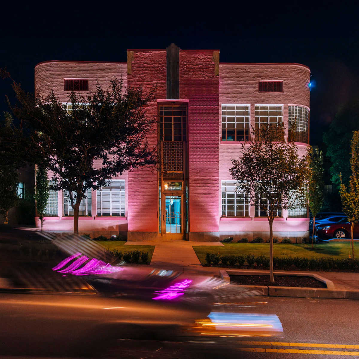 A pink-lit building with large windows stands at night, surrounded by trees and parked cars, while a car drives by on the street.