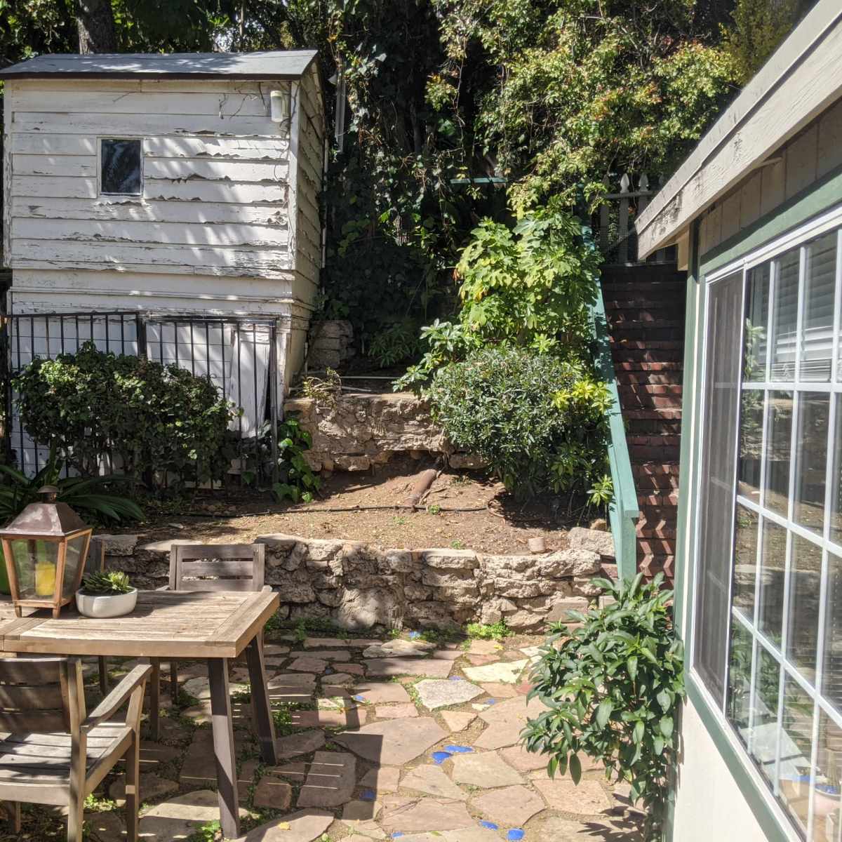 A small outdoor area with a wooden table and chairs, a weathered shed, a stone wall, and stairs leading up to a higher level.