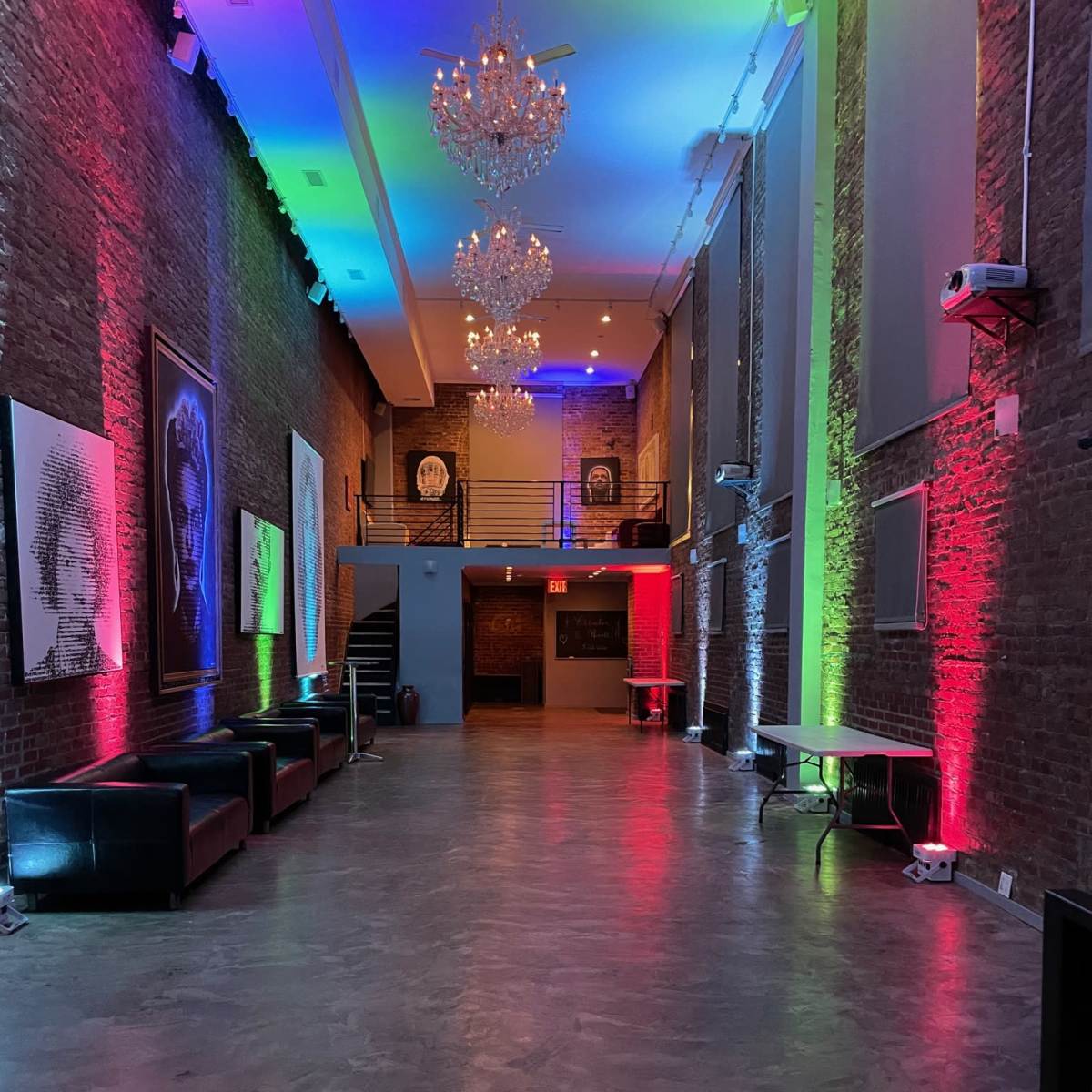 A spacious event hall featuring a polished concrete floor, brick walls, and vibrant colored lighting with chandeliers hanging from the ceiling.