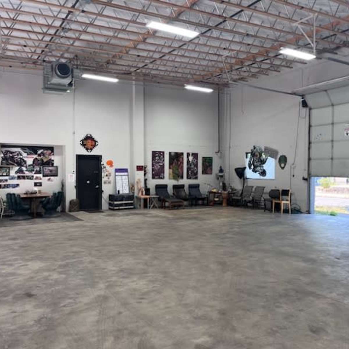 A spacious garage or workshop with unoccupied tables and chairs, wall art, and a large door that opens to an outdoor area.