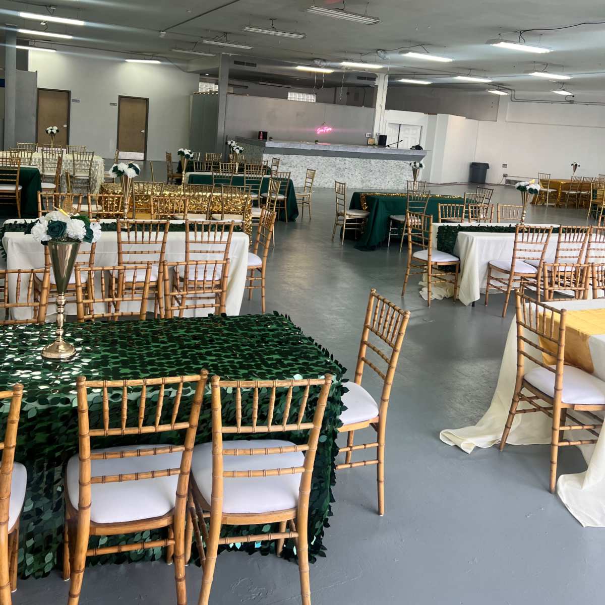 A spacious banquet hall arranged with multiple tables covered in various colored tablecloths and set with chairs facing a serving area.