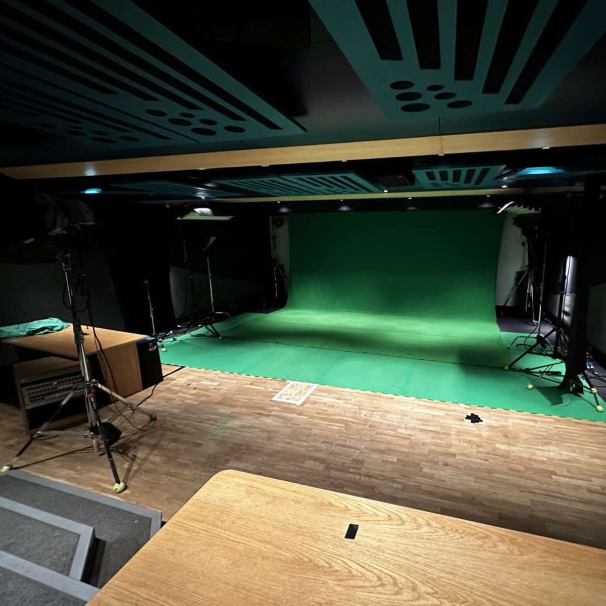 A green screen studio with lighting equipment set up around a green backdrop and a wooden table in the foreground.