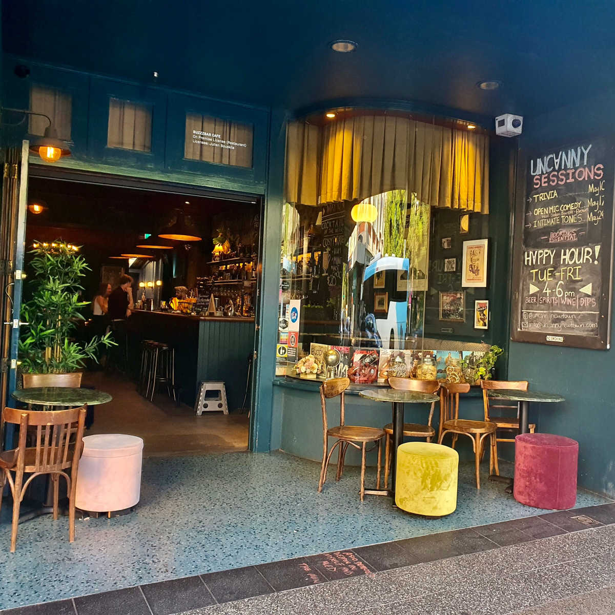The entrance of a bar with colorful stools and a chalkboard menu, featuring indoor seating and plants visible through the open doorway.