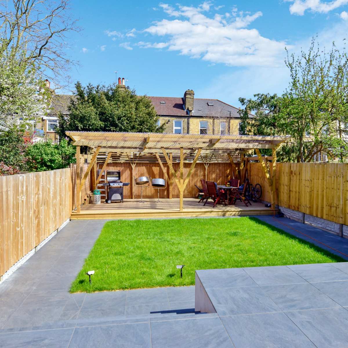 A well-maintained backyard featuring green grass, a wooden pergola with seating, and a fenced area surrounded by trees and a house in the background.