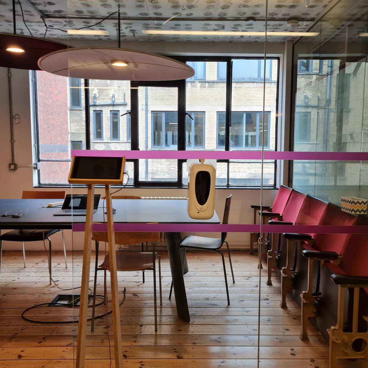 A conference room features a large table, chairs, and a modern device on a stand, with large windows providing natural light.