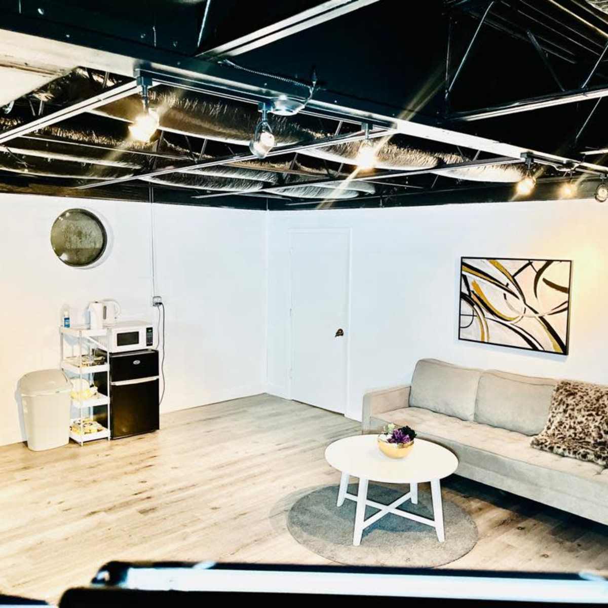 A modern room features a light gray sofa, a round coffee table with a floral centerpiece, a microwave, a small black refrigerator, and a round wall mirror against a white backdrop.