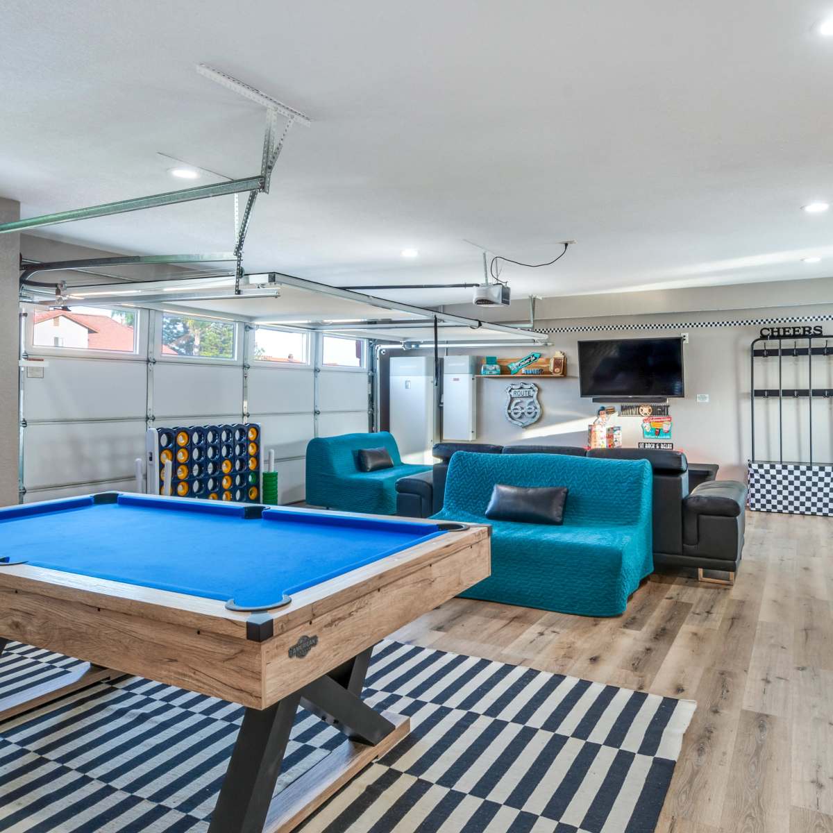 A spacious game room featuring a blue pool table, a seating area with teal furniture, a jukebox, and a garage door that opens to the outside.