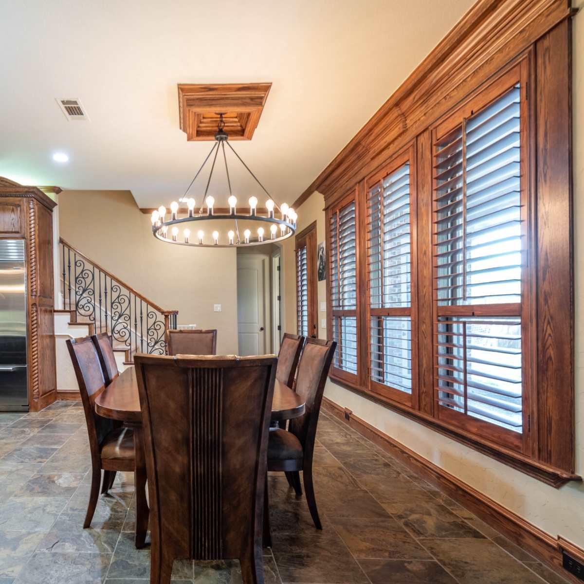 A modern dining area features a wooden table surrounded by chairs, illuminated by a large chandelier, and is adjacent to a kitchen with dark cabinetry and a staircase.