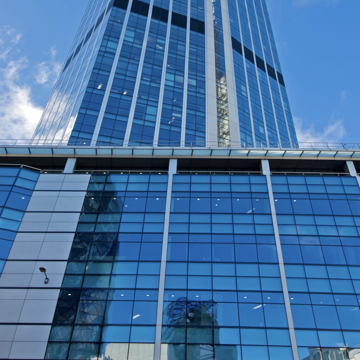 A tall, modern glass skyscraper reflects the sky and surrounding buildings, with a clear blue backdrop.