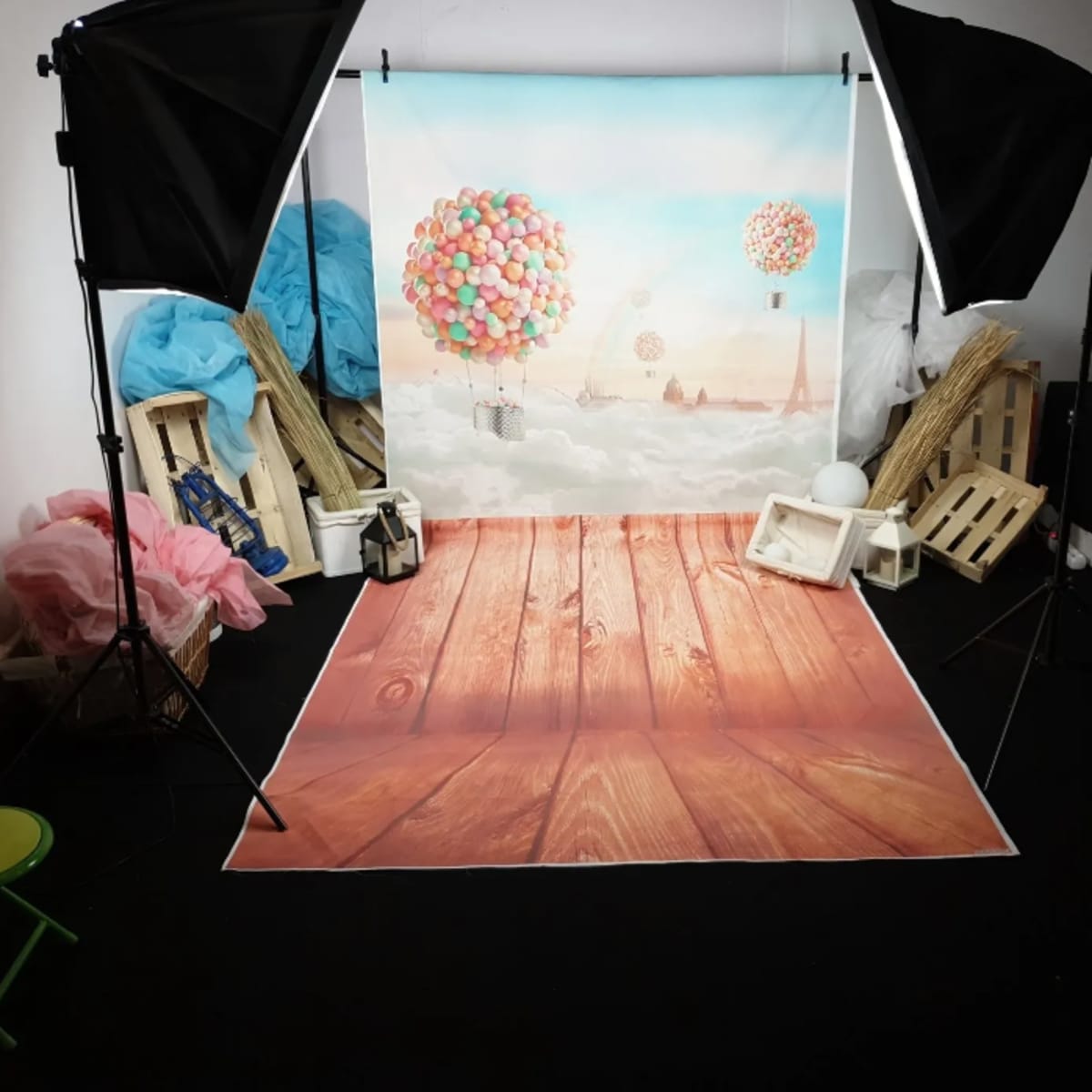 A photo studio setup features a backdrop of colorful balloons floating over a cityscape, with wooden floor props and studio lighting equipment.