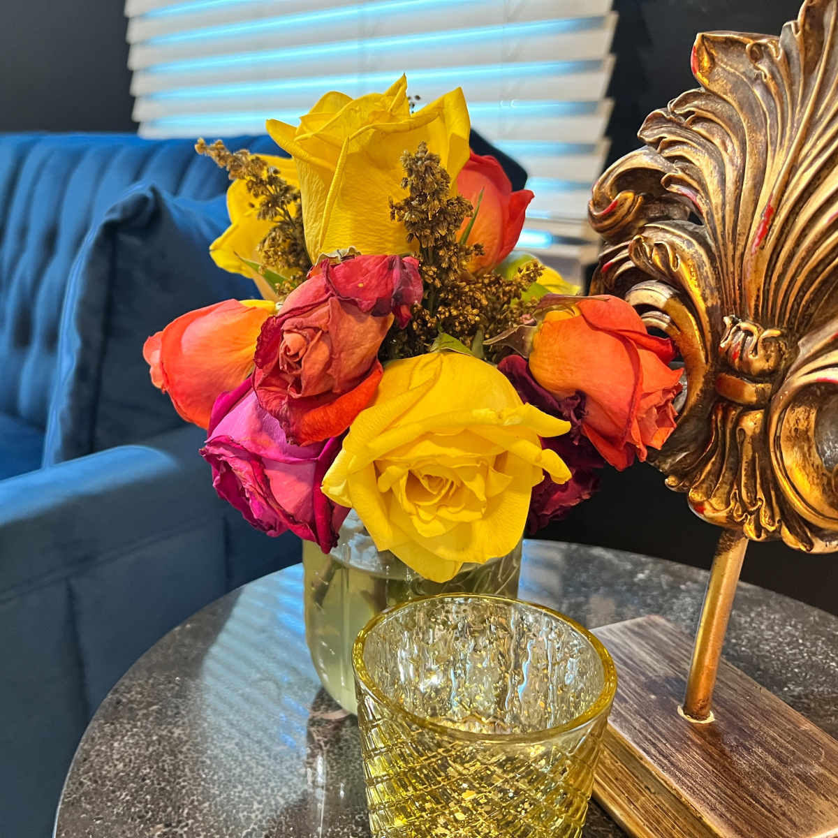 A bouquet of multicolored roses sits on a glass table next to a decorative gold item, with a blue sofa in the background.