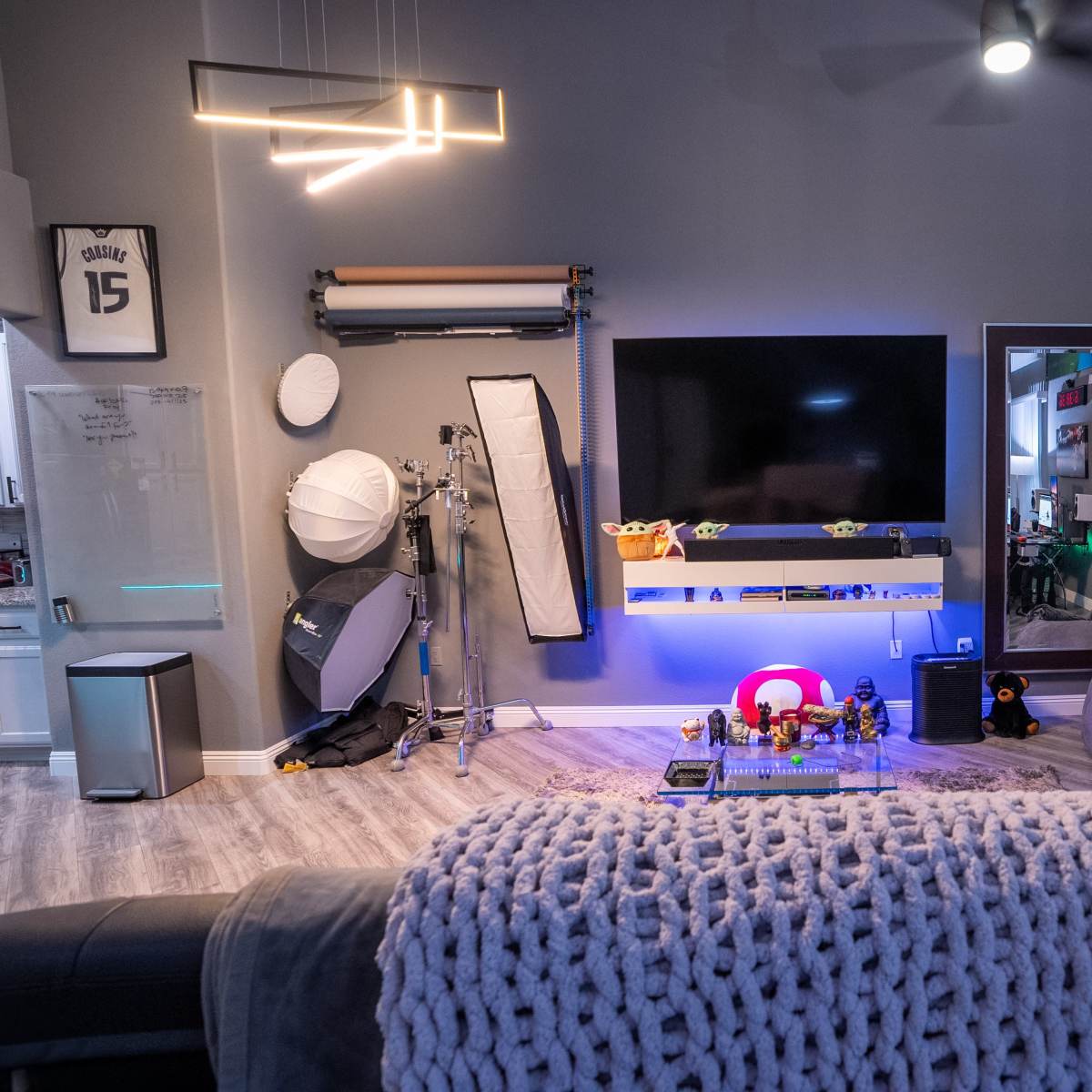 A modern living room with a large wall-mounted television, a cozy gray sofa, and various items including lighting equipment and decorative figures on display.