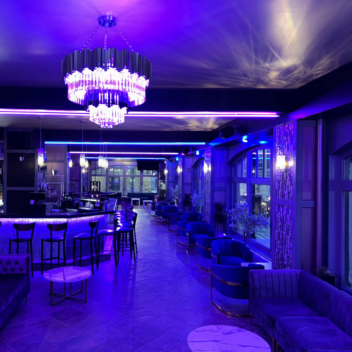 A modern lounge features plush seating, a long bar illuminated by blue and purple lights, and a large chandelier overhead.