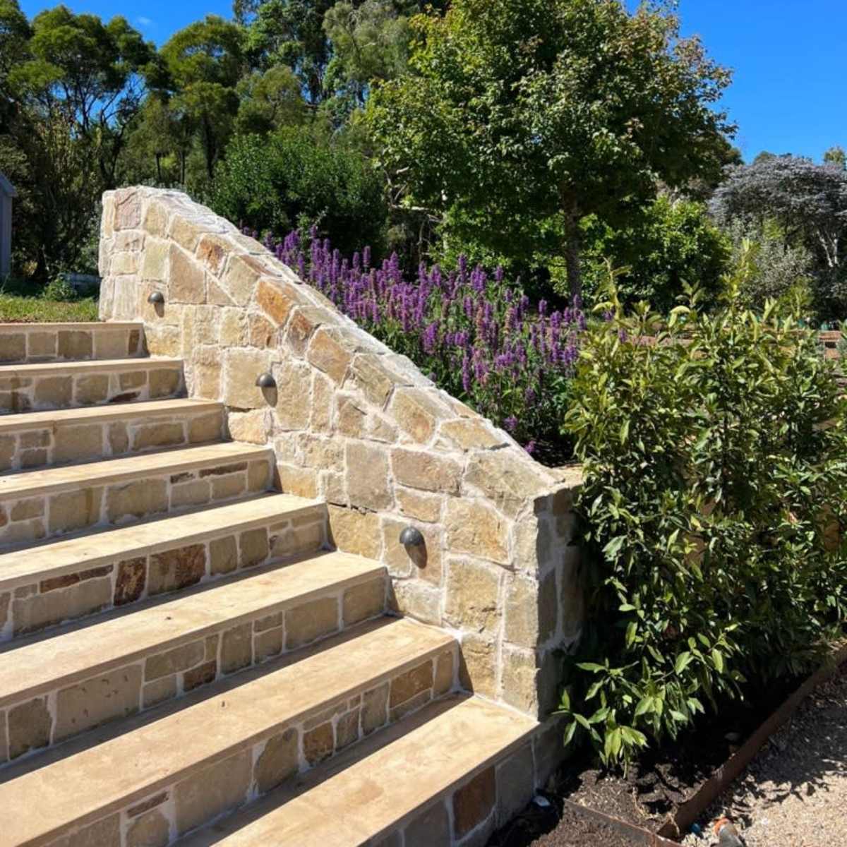 A stone staircase leading up to a garden with greenery and purple flowers under a clear blue sky.