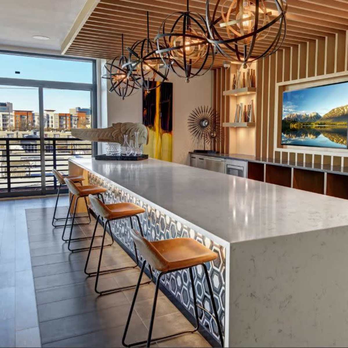 A modern kitchen with a long white countertop, beige bar stools, and decorative lighting fixtures above.