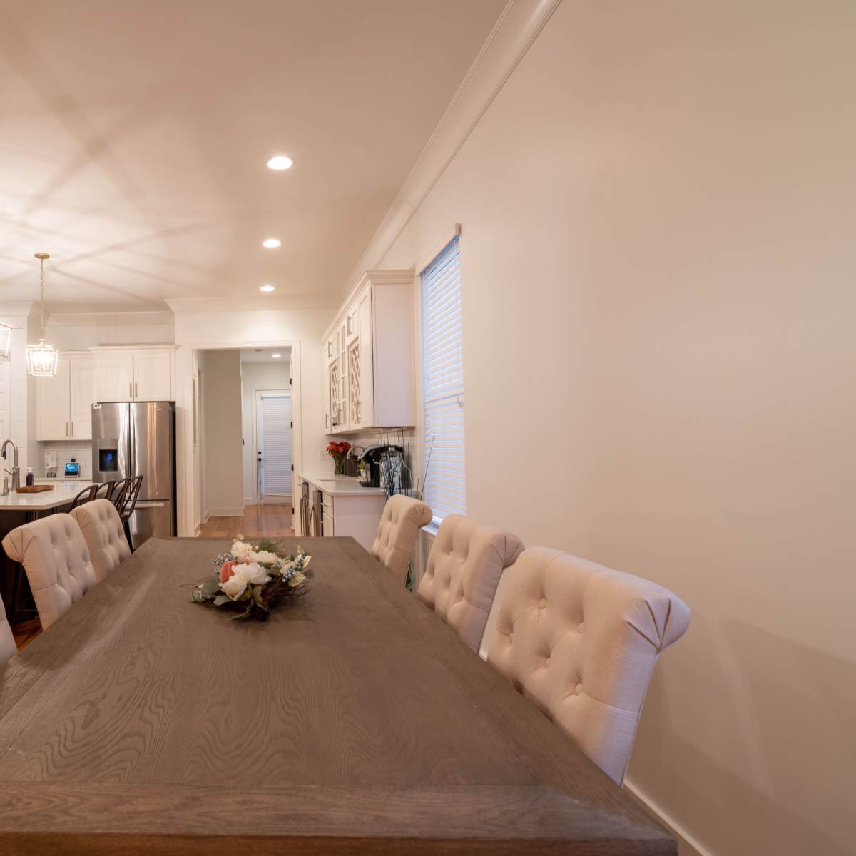 A spacious kitchen and dining area features a large wooden table surrounded by upholstered chairs, with modern appliances and cabinetry in the background.