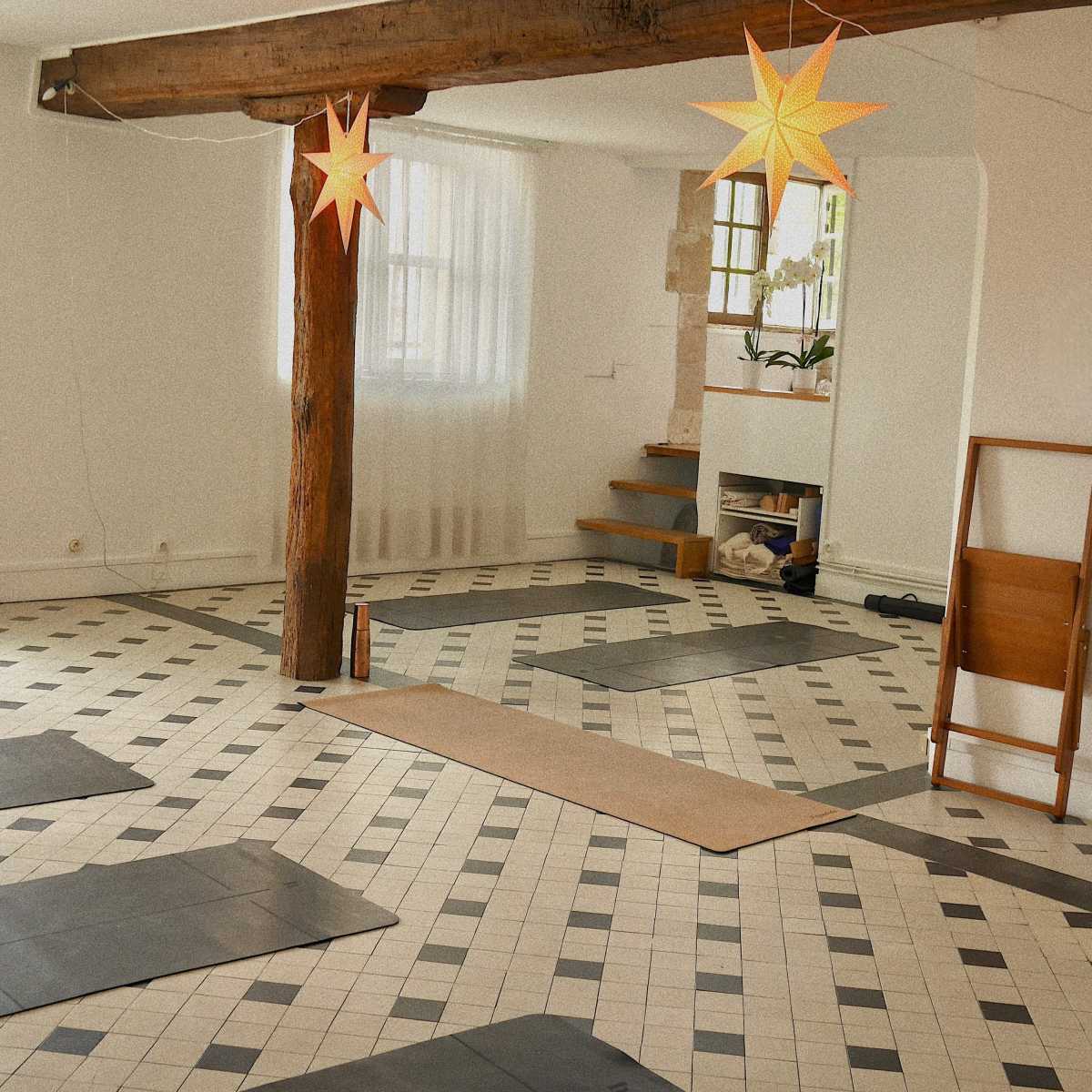 A yoga studio with black mats arranged on a tiled floor, featuring wooden beams and star-shaped paper lanterns hanging from the ceiling.