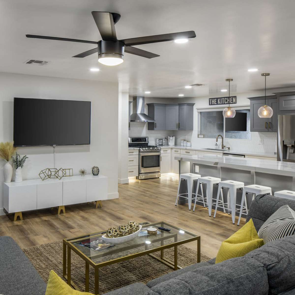 A modern living room and kitchen area with a ceiling fan, large TV, comfortable seating, and a sleek kitchen island with bar stools.