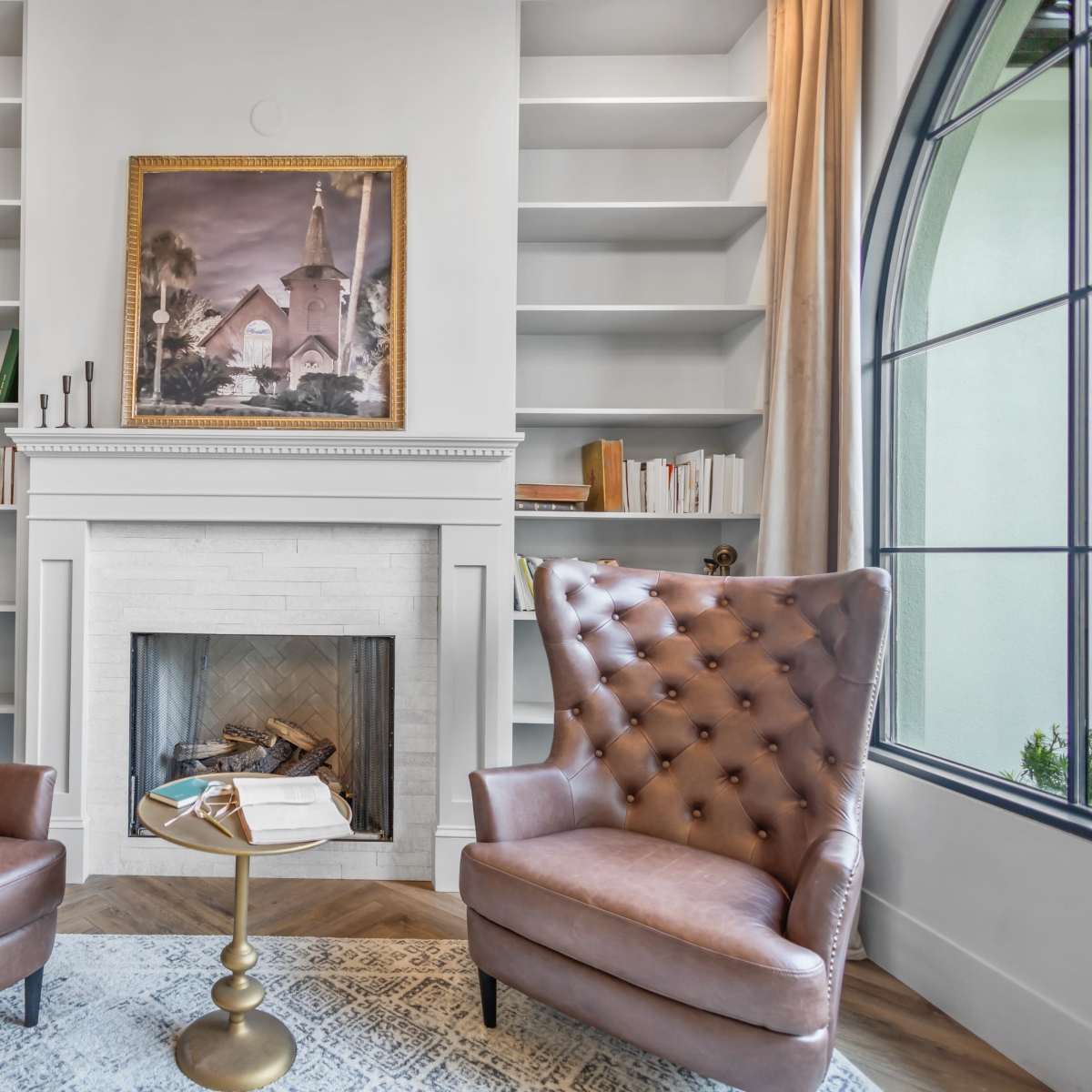 A cozy living space with two tufted brown armchairs facing a small round table, a fireplace surrounded by shelves, and a large window.