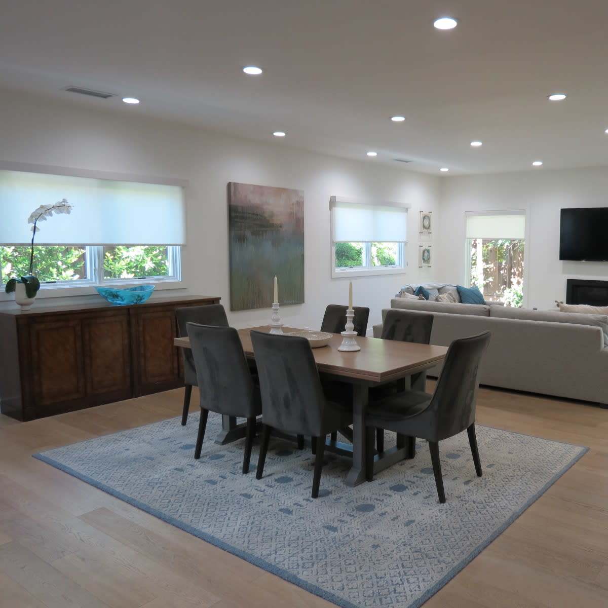 A modern living and dining area featuring a table with six chairs, a gray sofa, a television on the wall, and large windows with blinds letting in natural light.